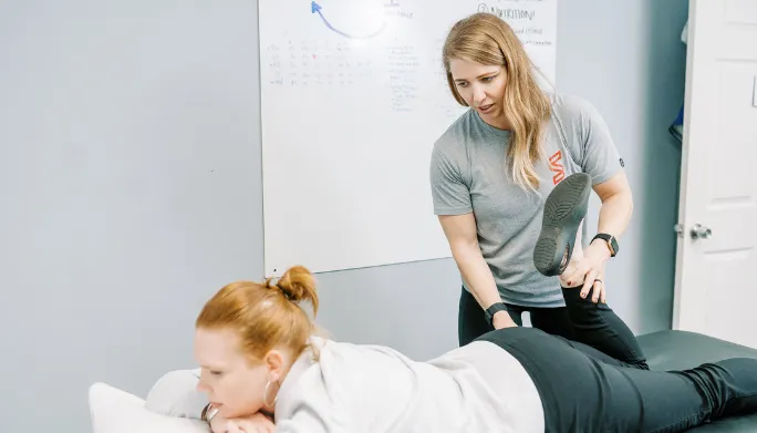 A therapist performing a hands-on hip and leg stretch on a patient lying face-down on a treatment table